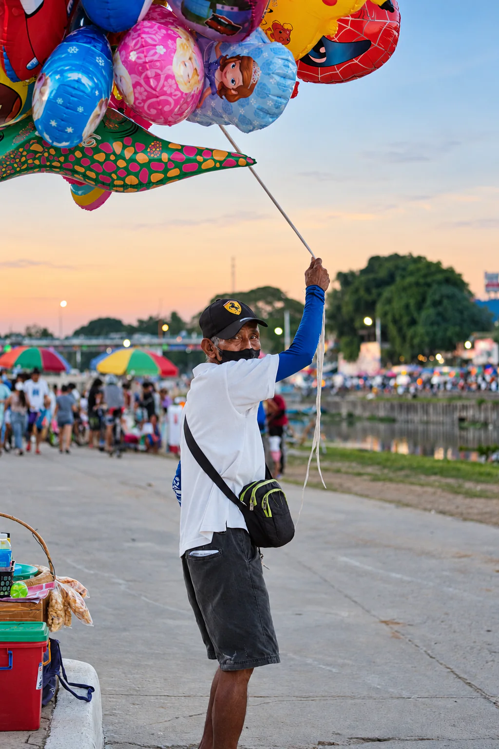 A balloon vendor at a crowded riverside fair looks back toward the camera while holding a long pole attached to a cluster of colorful character balloons overhead, with the setting sun glowing behind him.